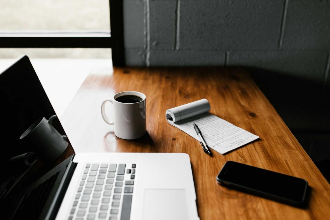 a laptop, a smartphone, a mug with coffee and a notebook with a pen are on a wooden desk