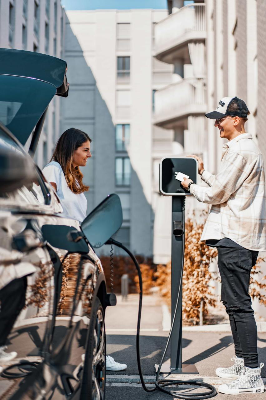 2 people are face to face in front of a car charging station. The person at the right is plugin in the cable. They are both smiling.