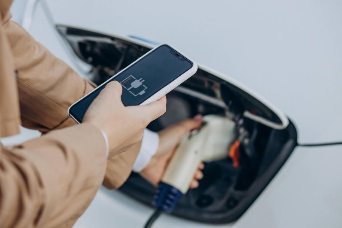 on the first layer of the picture, we can see a person holding a phone with its right hand. The screen shows a battery loading. With its left hand, the person is holding a charging cable for an electric car 